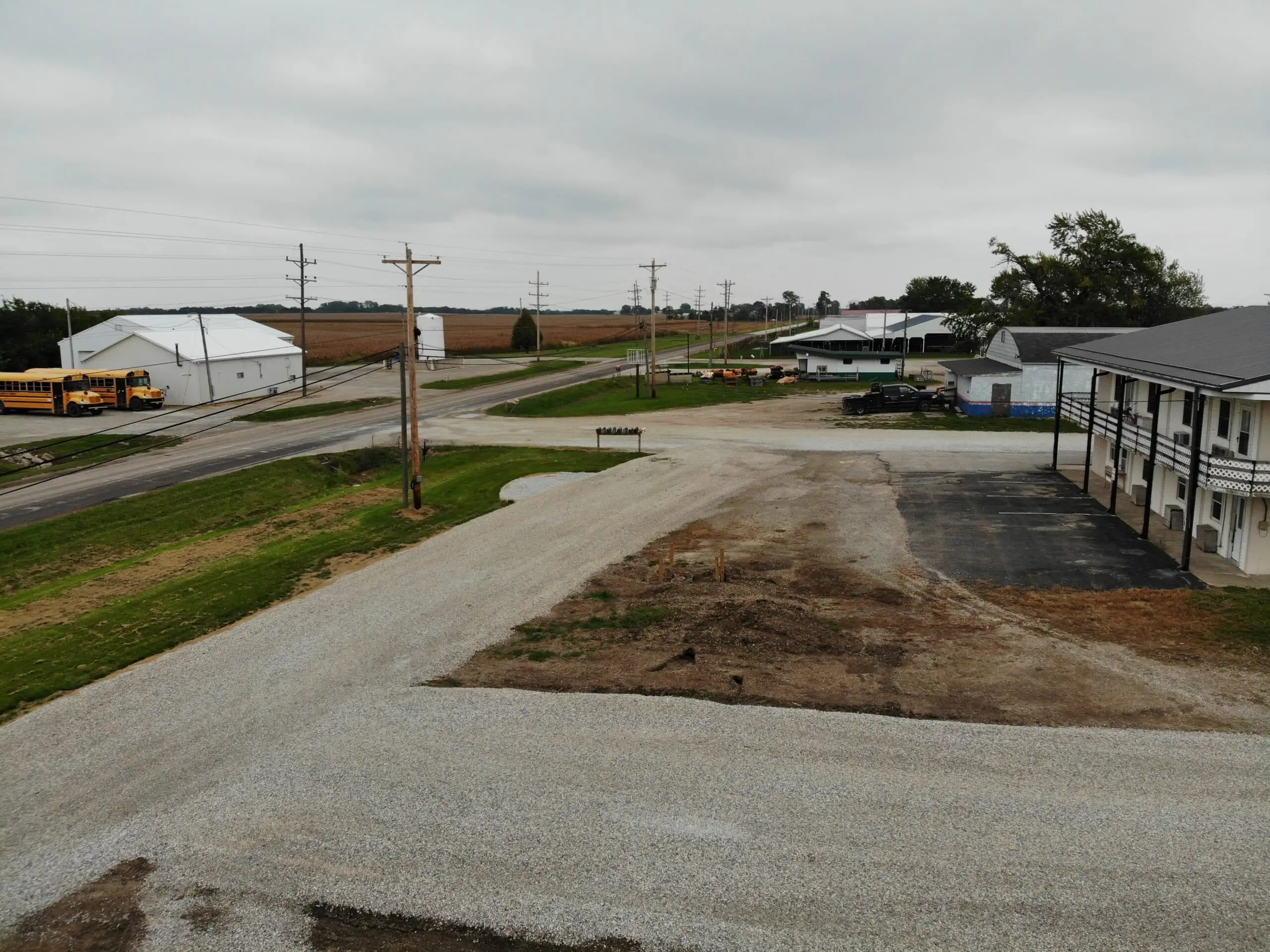 A rural scene with overcast skies, featuring a gravel road and parking area. A yellow school bus is parked near an intersection, surrounded by fields, power lines, and farm buildings. A two-story building is visible on the right.