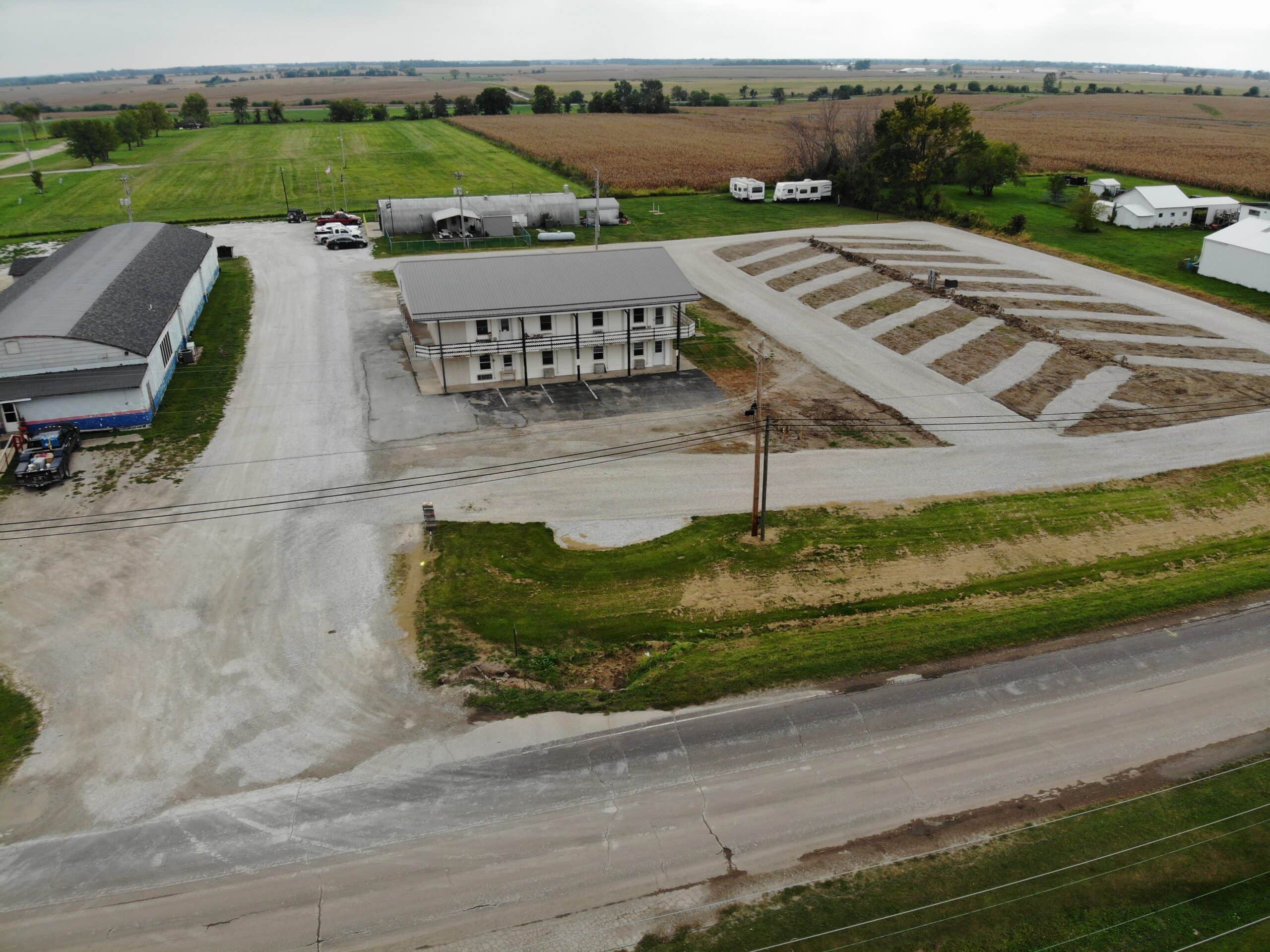 An aerial view of a rural area with a large building in the center, surrounded by gravel lots and fields. Farm buildings and vehicles are visible in the background under a cloudy sky.