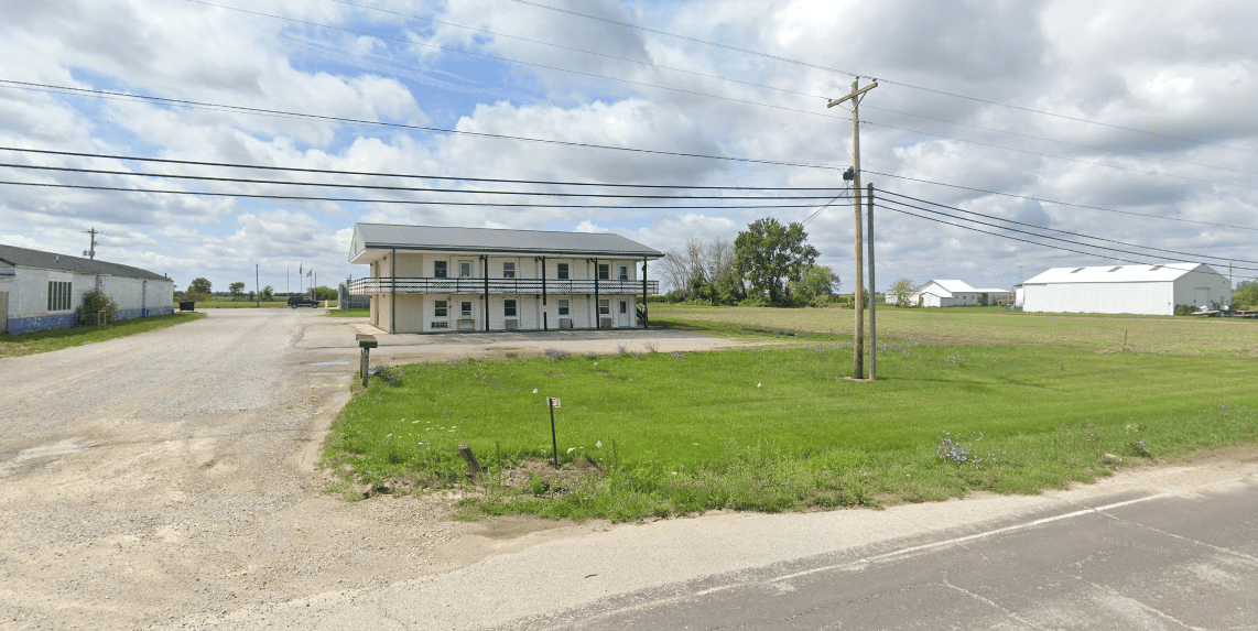 A two-story, beige building stands alongside a rural road. It's surrounded by green grass, a gravel driveway, and power lines. The sky is partly cloudy, and there are additional buildings in the background.